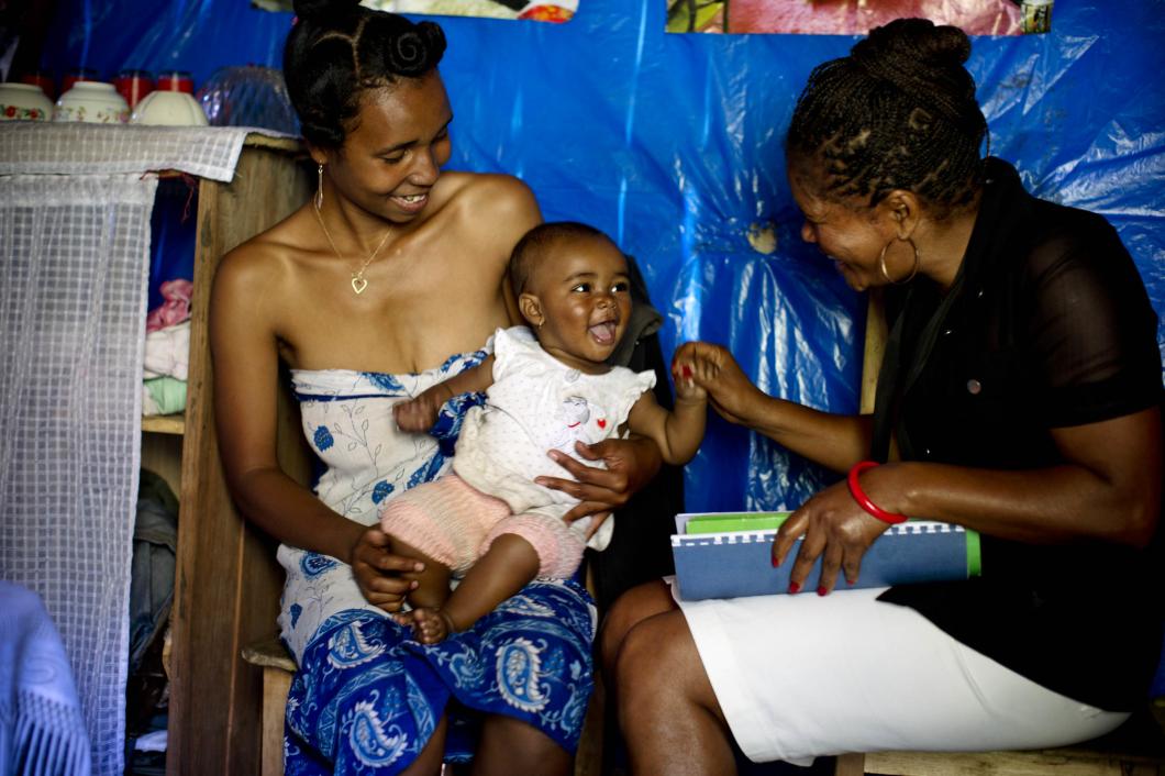 A woman holds her infant while a nurse smiles and holds her hand.